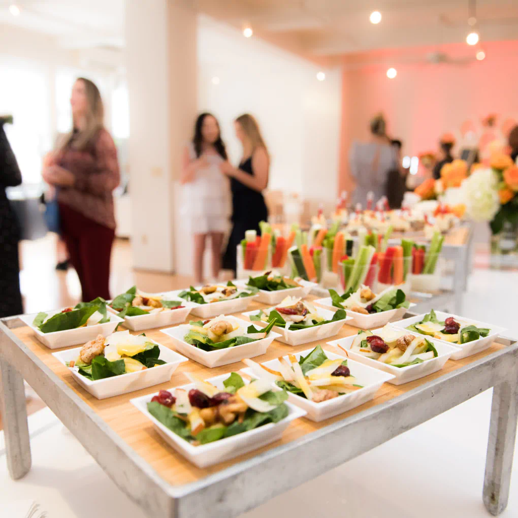 A large spread of salads and vegetables sit on a serving table, while groups of people converse in the background.