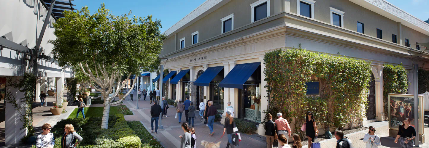People walk the sidewalks of Biltmore Fashion Park. One woman sits on the edge of a fountain. The many storefronts are seen in the backdrop.