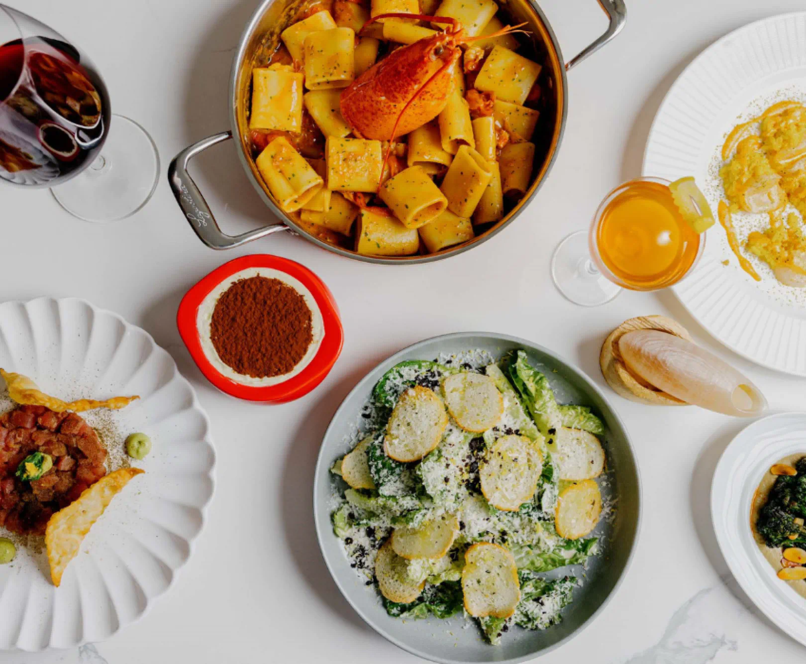 A spread of lobster pasta and caesar salad sits on a white table. It is served with white and red wine.