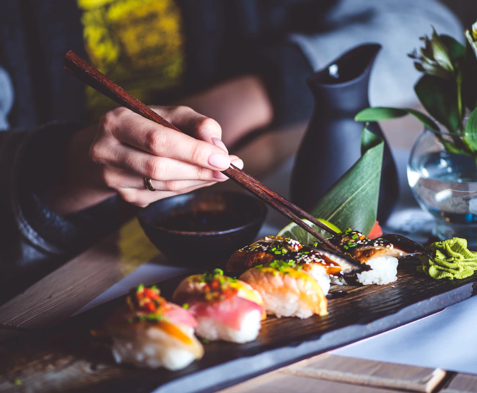 A person grabs a piece of sushi served on a black board with wooden chopsticks.