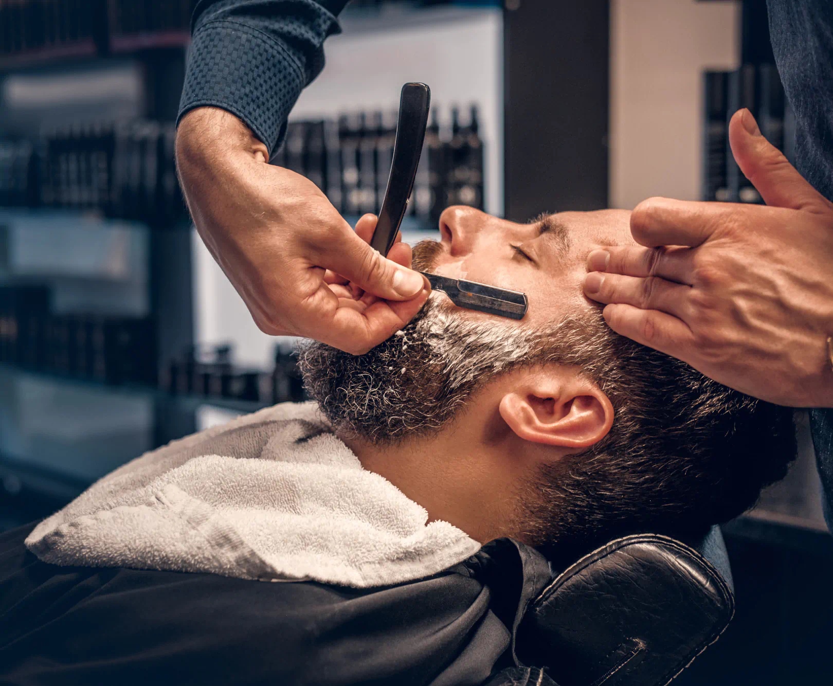 A bearded man sits in a barber's chair as he get a shave with a straight razor.