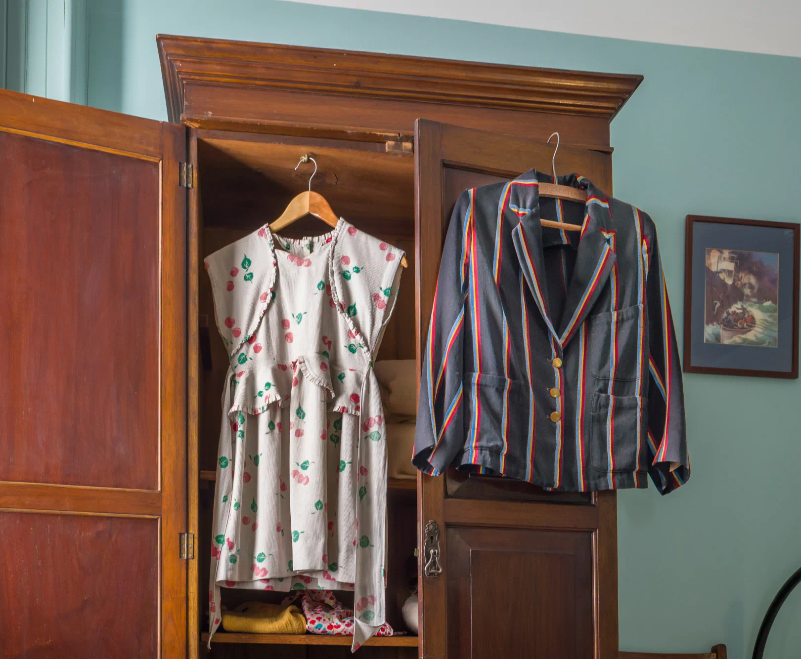 Two pieces of clothing hang from a cherry wood armoire. One is a white floral dress and the other is a dark gray suit with colorful stripes.
