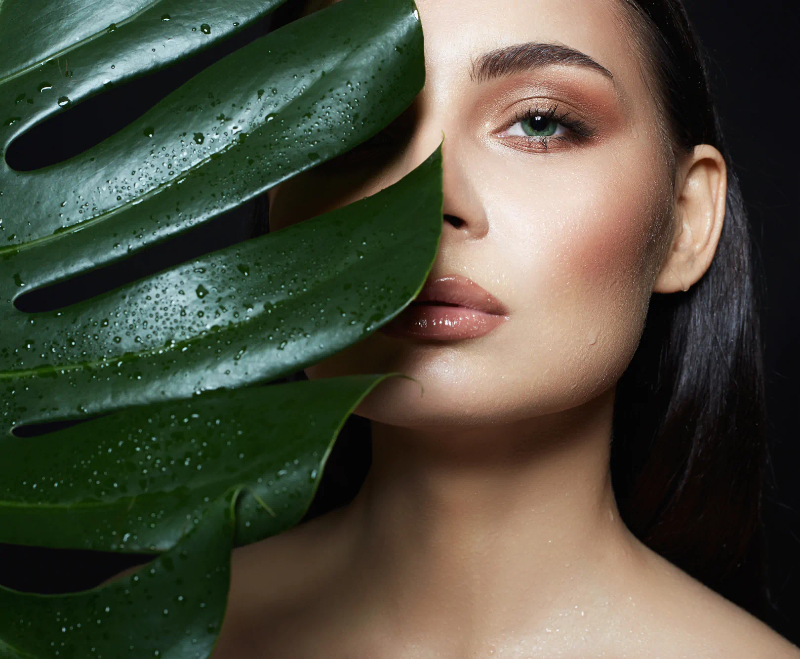 A girl with striking green eyes covers half of her face with a large green leaf.
