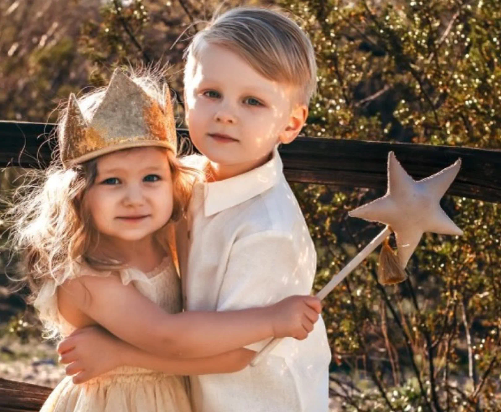 Two children hug in the outdoors. One girl wear a golden crown and carries a wand. The other boy wears a cream button down.