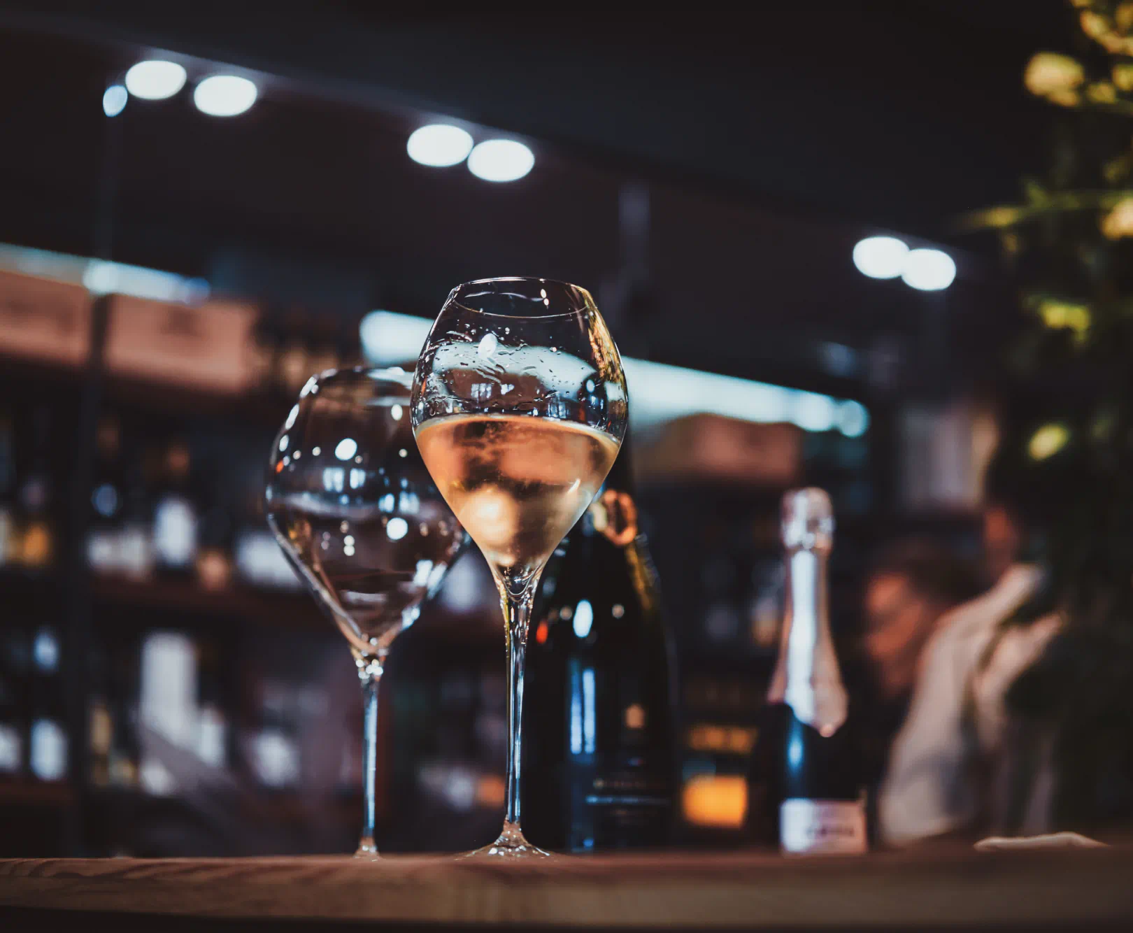 Two glasses sit on a dimly lit bar. One is filled with white wine.