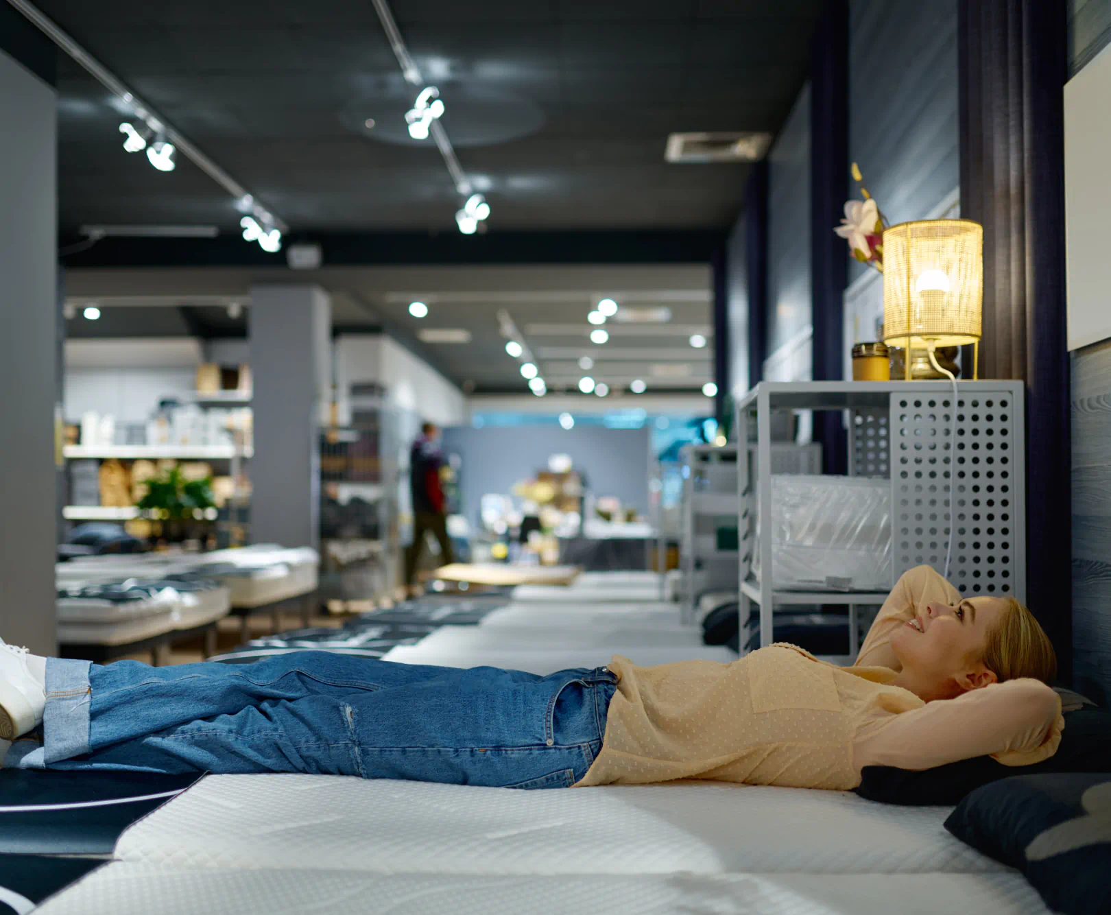 A woman smiles and she lays down on a showroom mattress.