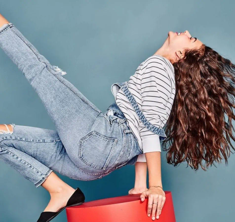 Joyful woman perched on a red cylinder dressed in jeans and a sweater, kicking her leg in the air with her head thrown back