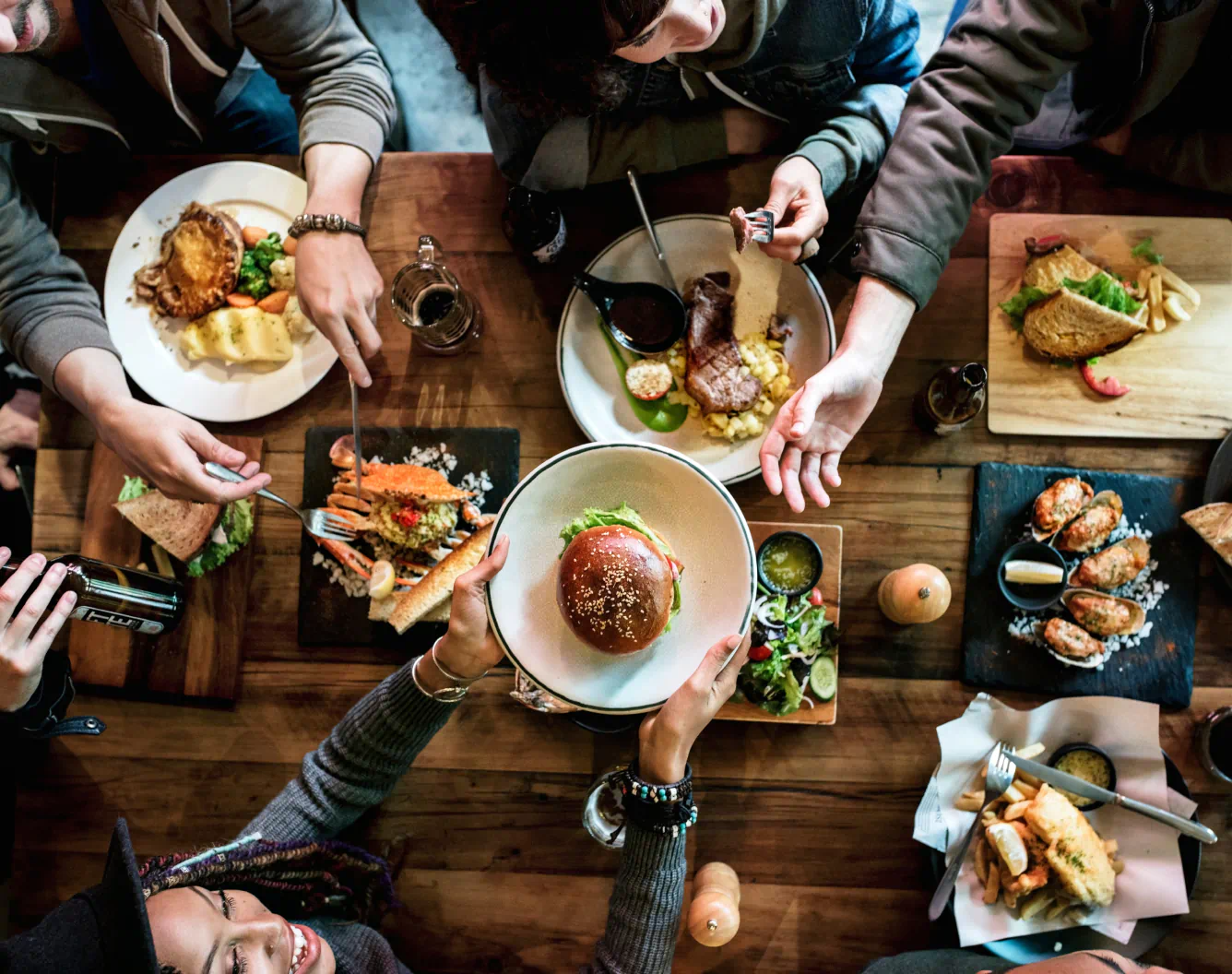 overhead view of a table laden with food including crab, salad, and oysters. One friend passes a burger to someone on the opposite side of the table.