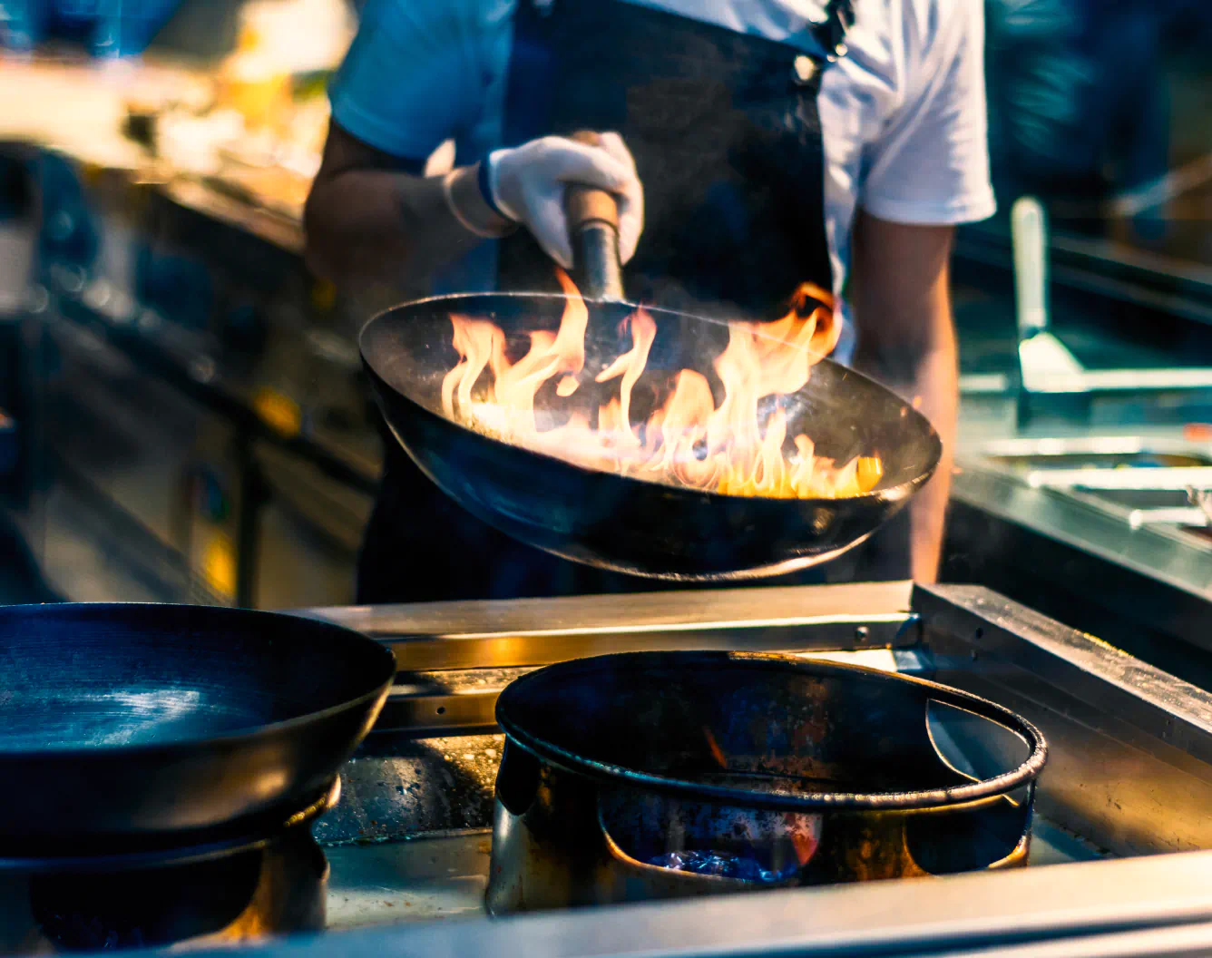 A wok with bright orange flames is held by a chef in a black apron in a fine restaurant.