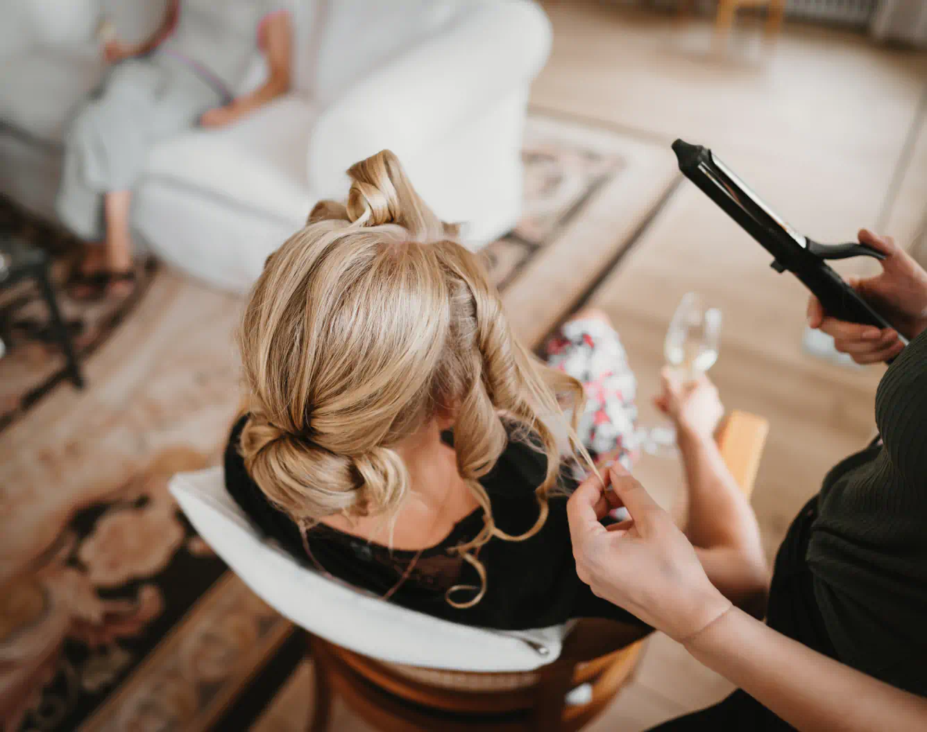 A hairdresser curls the blond hair of a person sitting and holding a wine glass