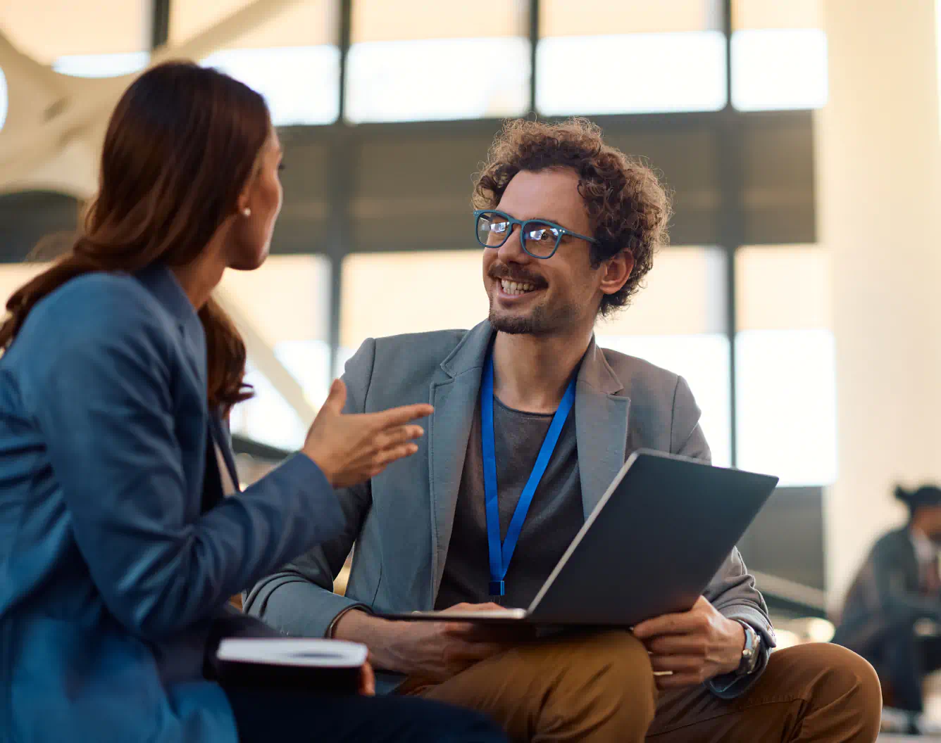 Two business professionals sit looking at each other, a laptop on one of their knees. They wear identifying lanyards and look engaged in inspiring conversation.