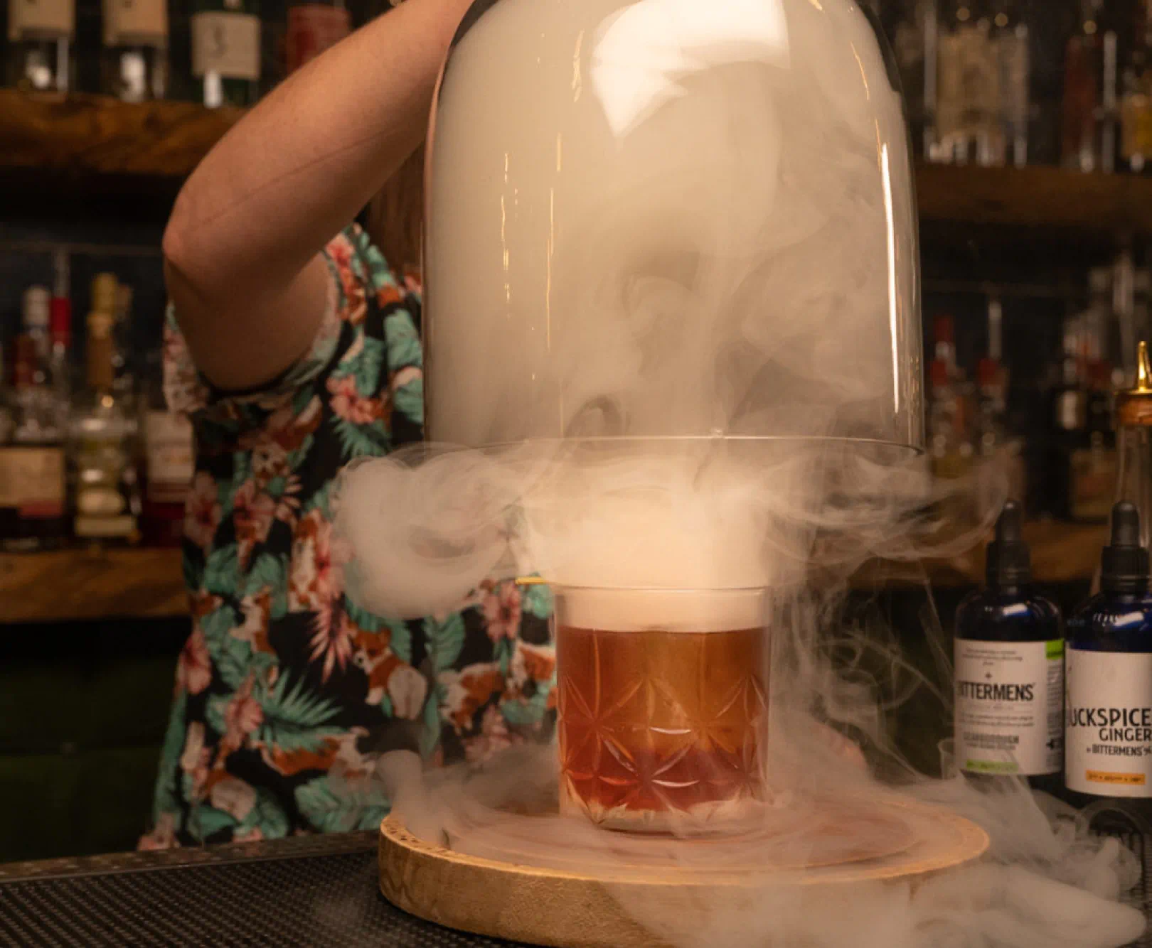 A bartender lifting a glass dome from a glass of bourbon with a cloud of smoke