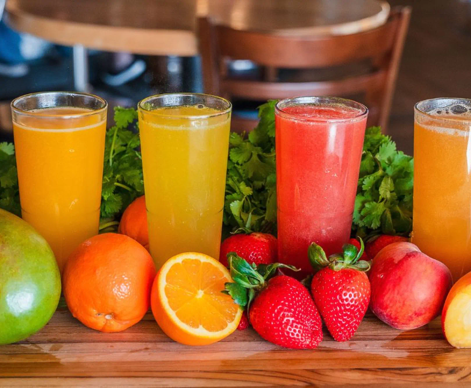 Four drinks, orange, yellow, red, and tangerine sit on a wooden surface surrounded by oranges, strawberries, and peaches.