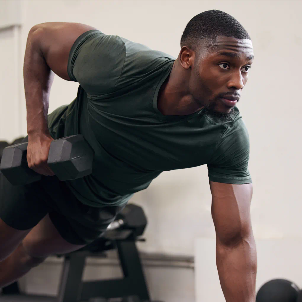 A strong man does a lateral row with dumbbells in had in a plank position in a gym.