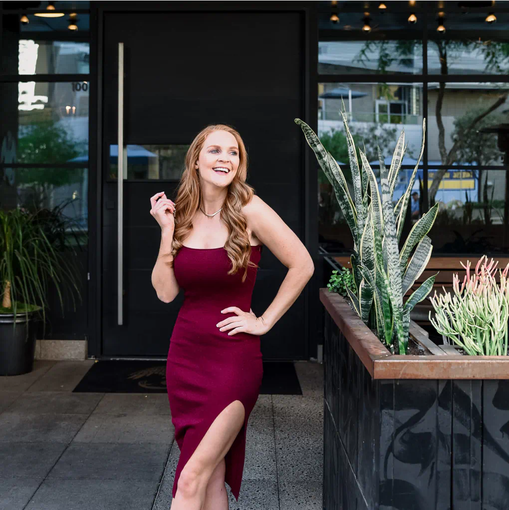 A smiling, fashionably dressed woman stands in front of a darkly colored restaurant in a red dress by some landscaping.