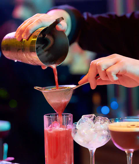 Close up of a bartender artfully pouring a red cocktail through a strainer into a glass on the bar. A nice glass with ice, and an espresso martini also sit on the bar.