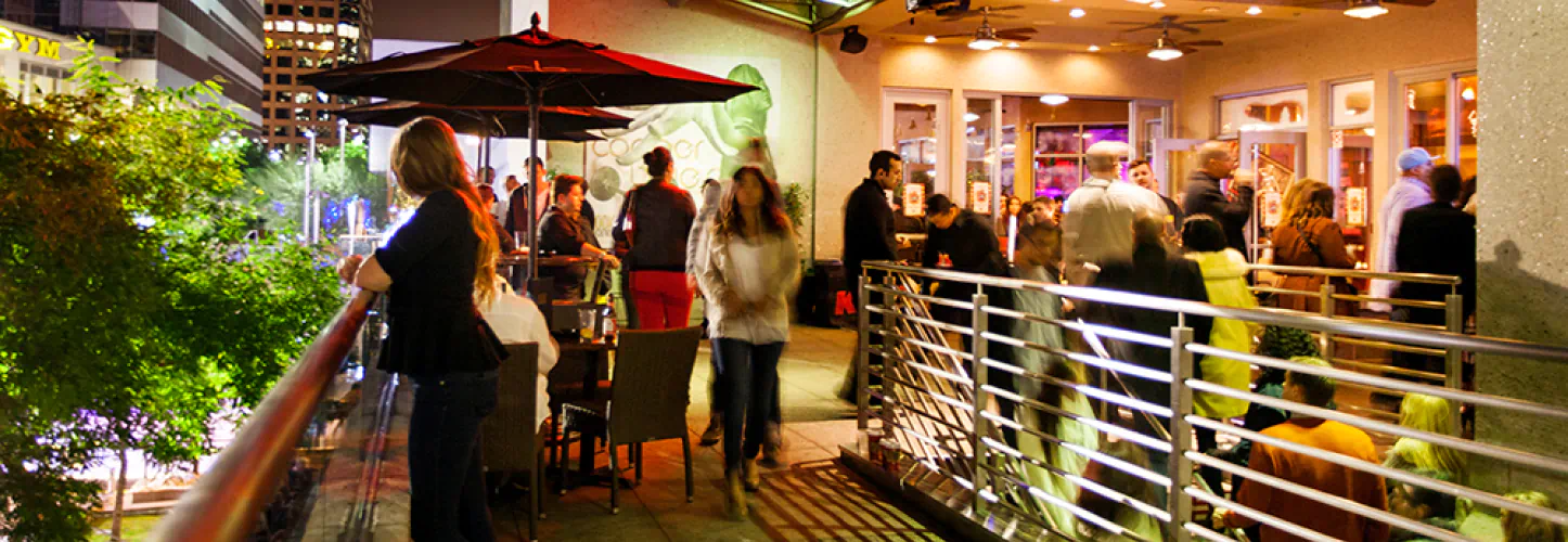 Long exposure shot of the exterior of a restaurant on the upper level. People walk up the stairs towards the restaurant and some are looking over the balcony toward the street below.
