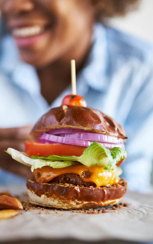 an image of a burger on a pretzel bun with a cherry tomato skewered on top, out of focus in the background sits a smiling woman.