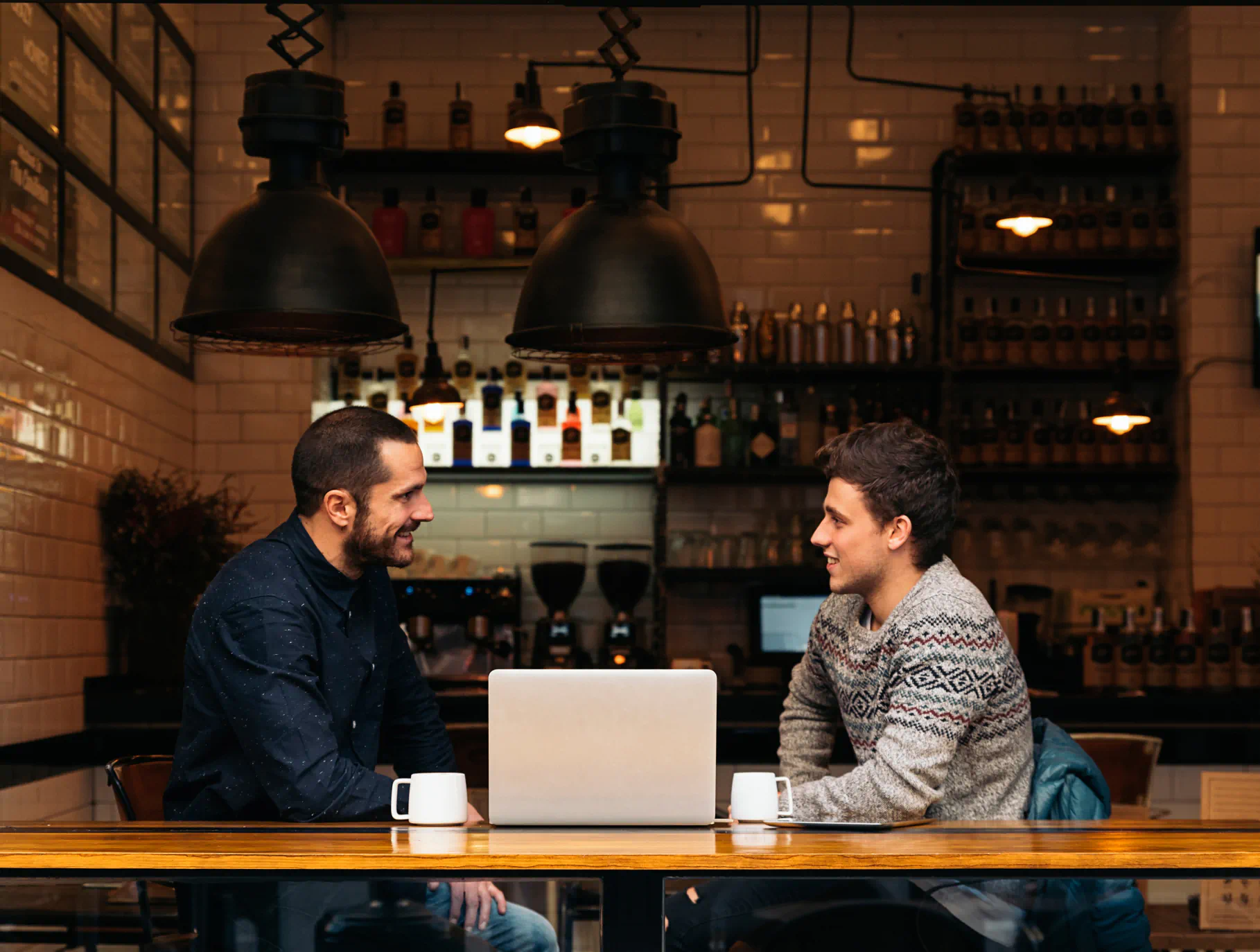 Two men sit at a table with a laptop and coffee mugs in a modern café with white tiled walls and industrial-style pendant lamps.
