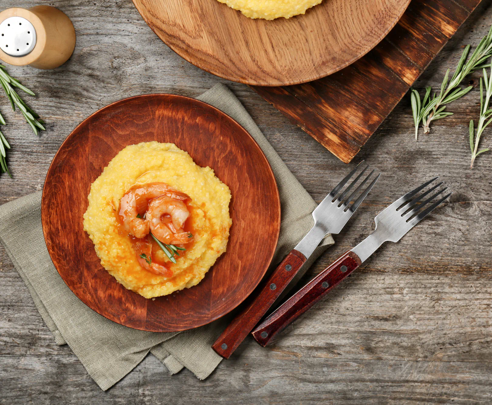 Wooden plate with shrimp over grits, placed on a grayish-green napkin, with two forks and sprigs of rosemary on a rustic wooden table.