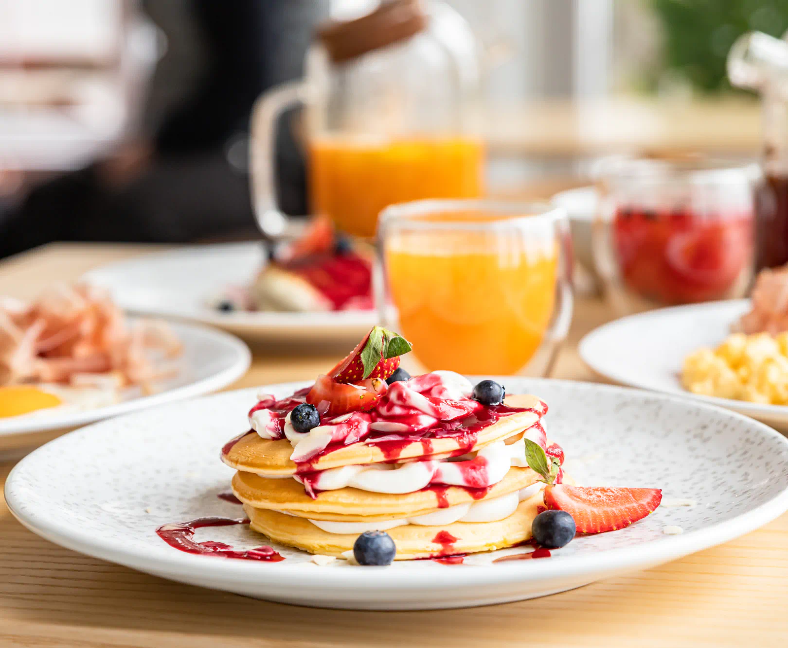 A stack of pancakes topped with strawberries, blueberries, mint, and drizzled with red and white sauce on a breakfast table.