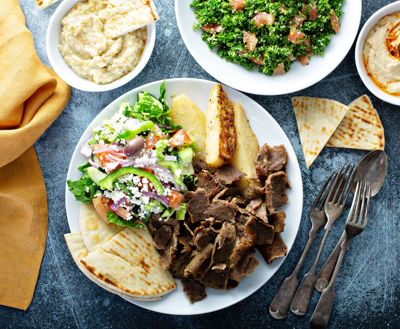 A plate of Mediterranean food with salad, roasted potatoes, gyro meat, and pita bread, accompanied by small bowls of hummus, baba ganoush, and tabbouleh.