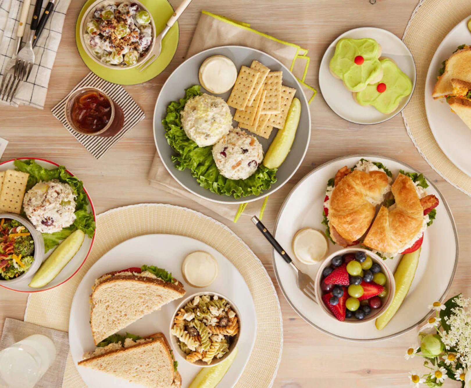 A tabletop with various plates of sandwiches, salads, crackers, pickles, and side dishes arranged in an aerial view.