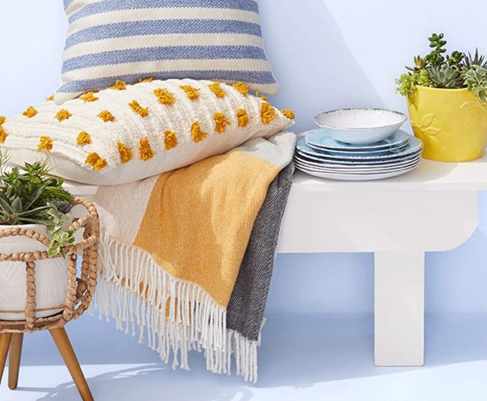 White bench with cushions, a blanket, stacked plates, and a yellow planter with succulents on top. A planter with trailing plants is on the floor next to the bench.
