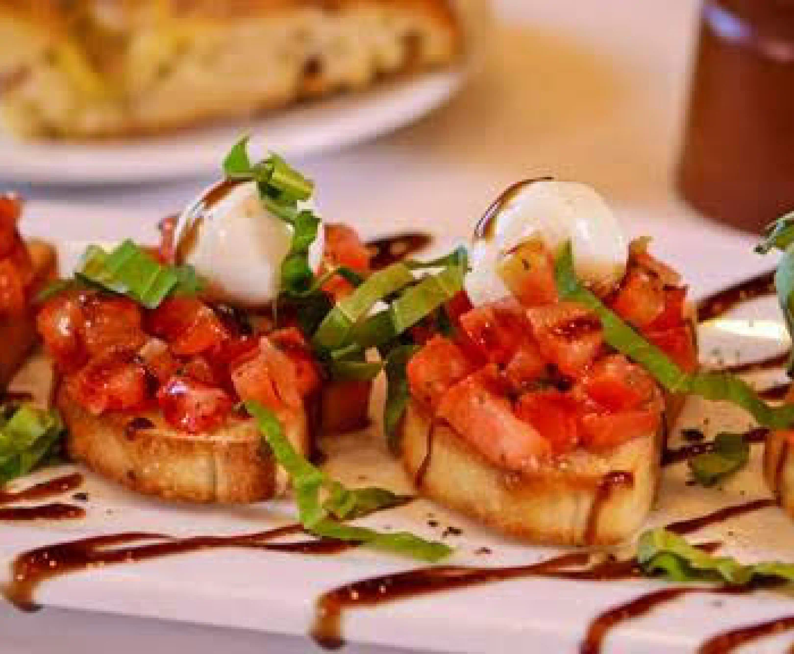 Three bruschetta with diced tomatoes, basil, and mozzarella on a white plate drizzled with balsamic glaze.