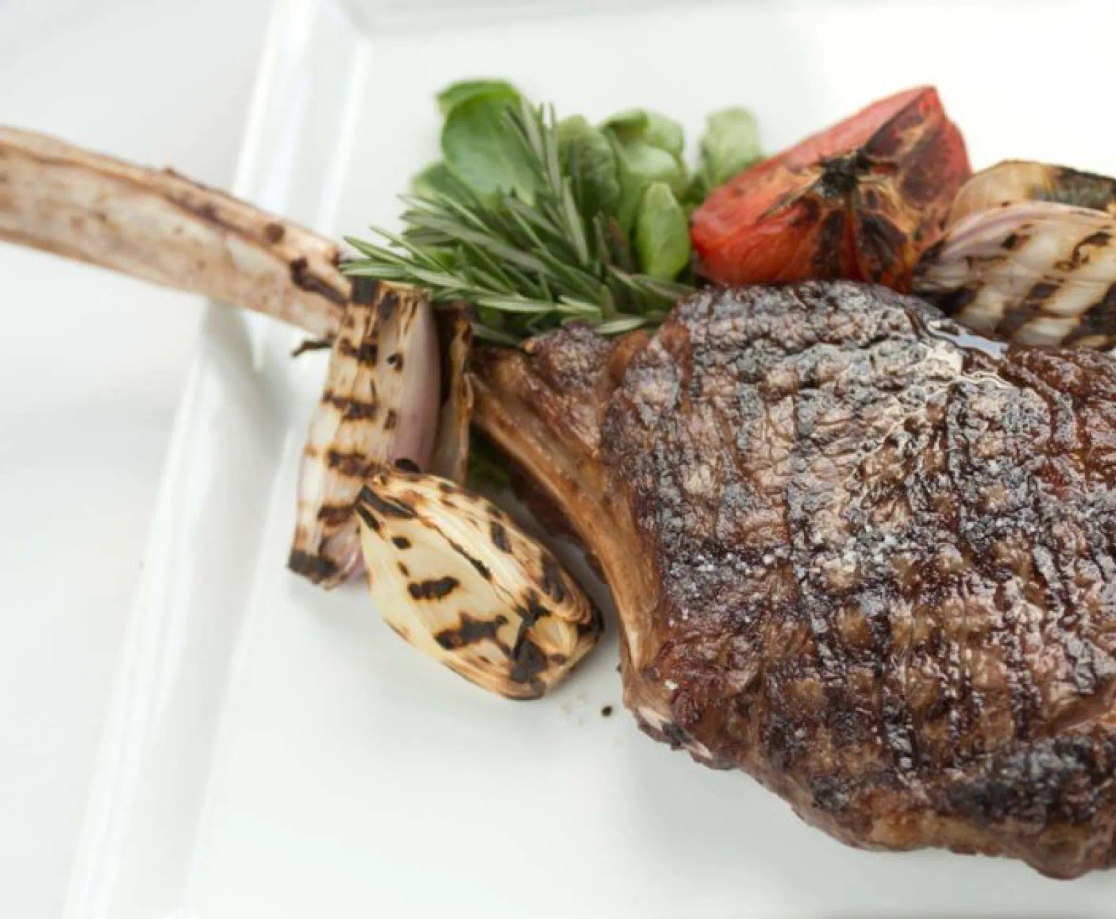 a tomahawk steak arranged on a square white plate and surrounded by roasted garlic cloves, tomatoes and various herbs