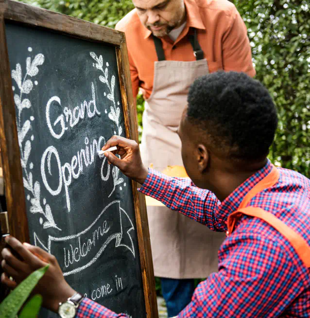 Two men in aprons kneel around a sandwich board sign, the first man writes on the sign in script "Grand Opening".