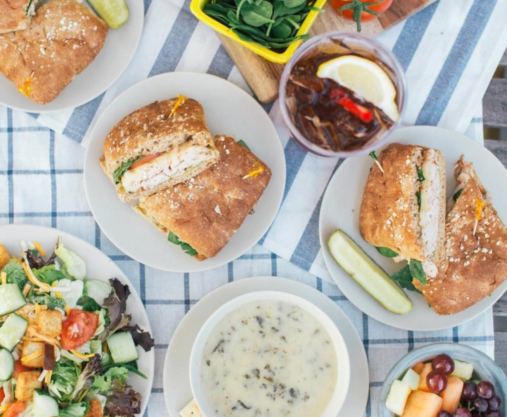 A table set with sandwiches, salad, soup, a drink, fresh spinach, and a bowl of fruit on a checkered tablecloth.