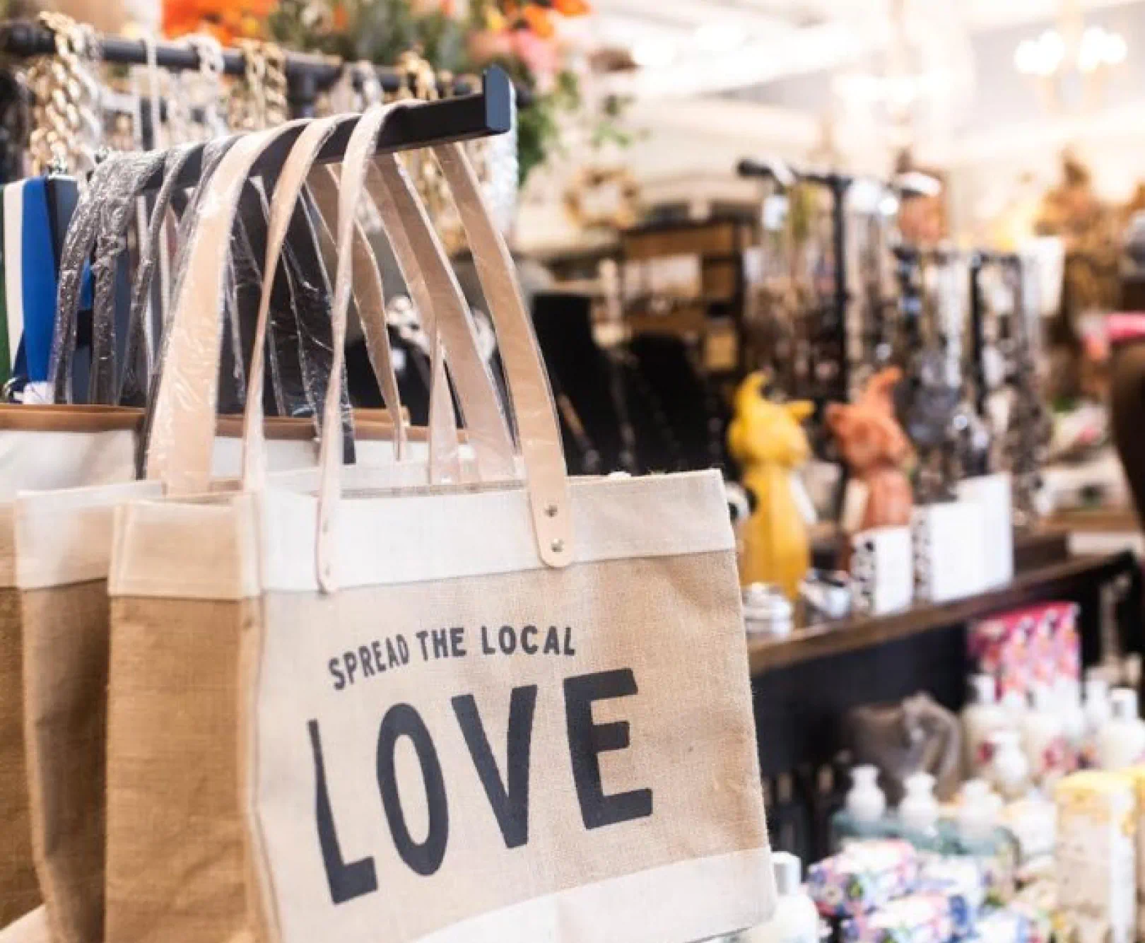 Canvas tote bags hanging on a rack in a shop, with the printed text "SPREAD THE LOCAL LOVE." Assorted items on shelves in the background.