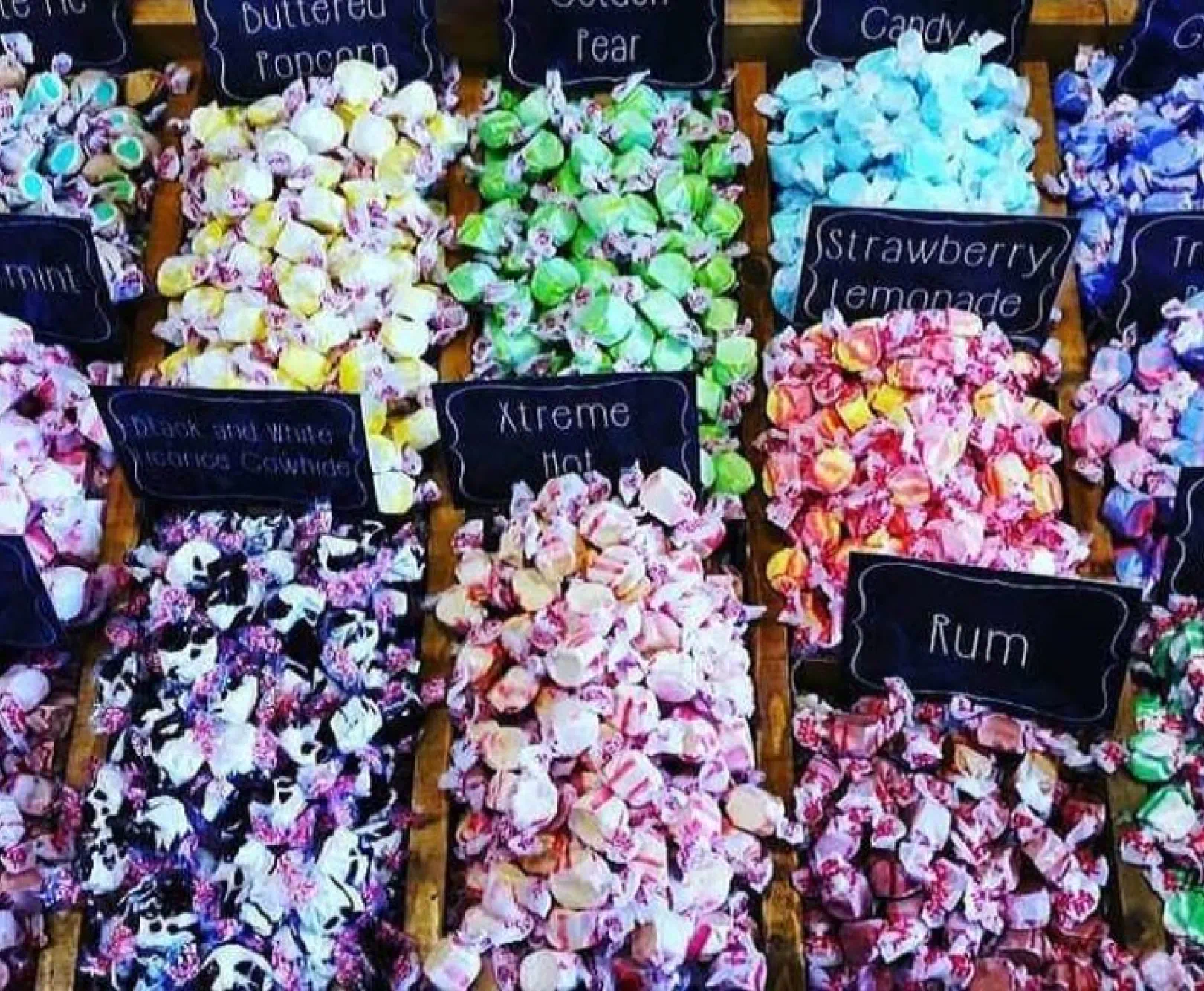various flavors of saltwater taffy arranged in wooden boxes with labels listing buttered popcorn, pear, min, strawberry lemonade, black and white locorice, xtreme hot, and rum
