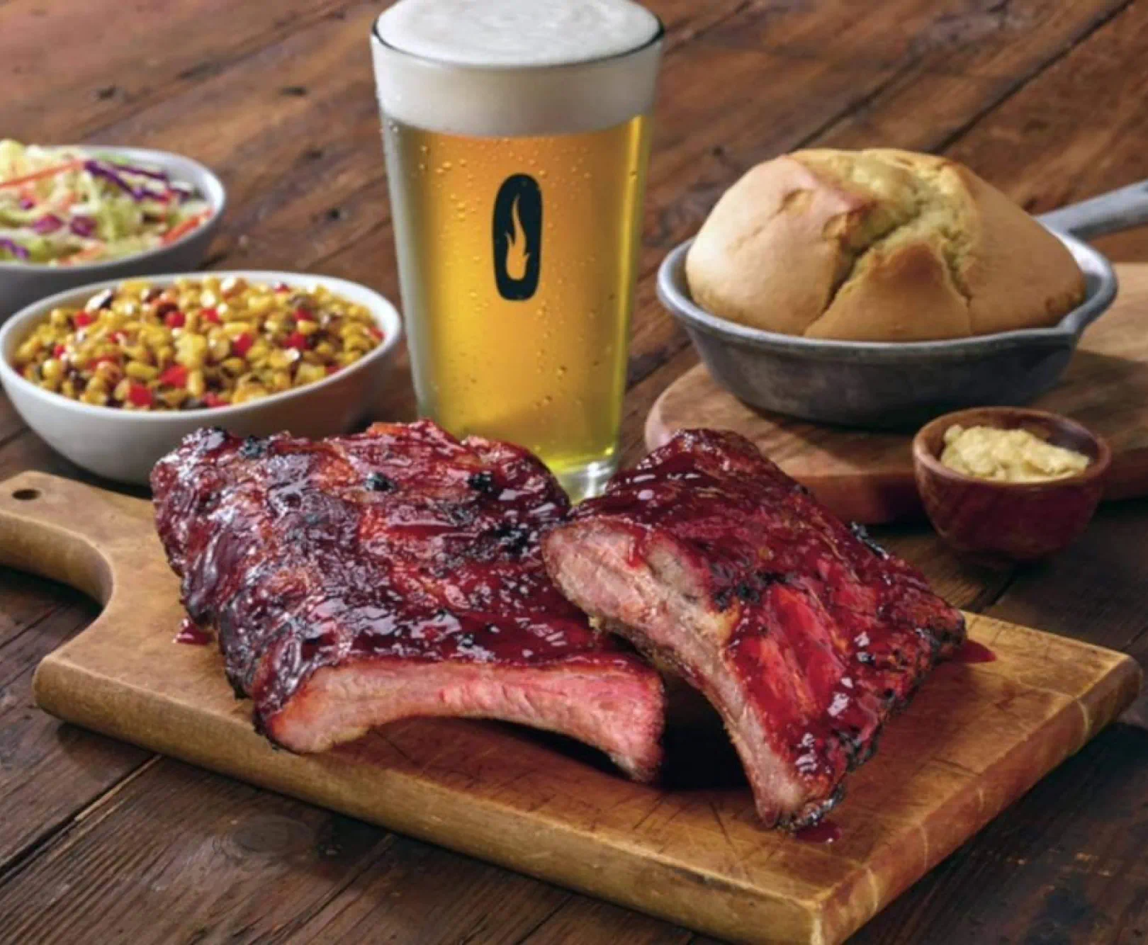 Barbecued pork ribs with a pint of beer, coleslaw, corn salad, bread, and a side of butter on a wooden surface.