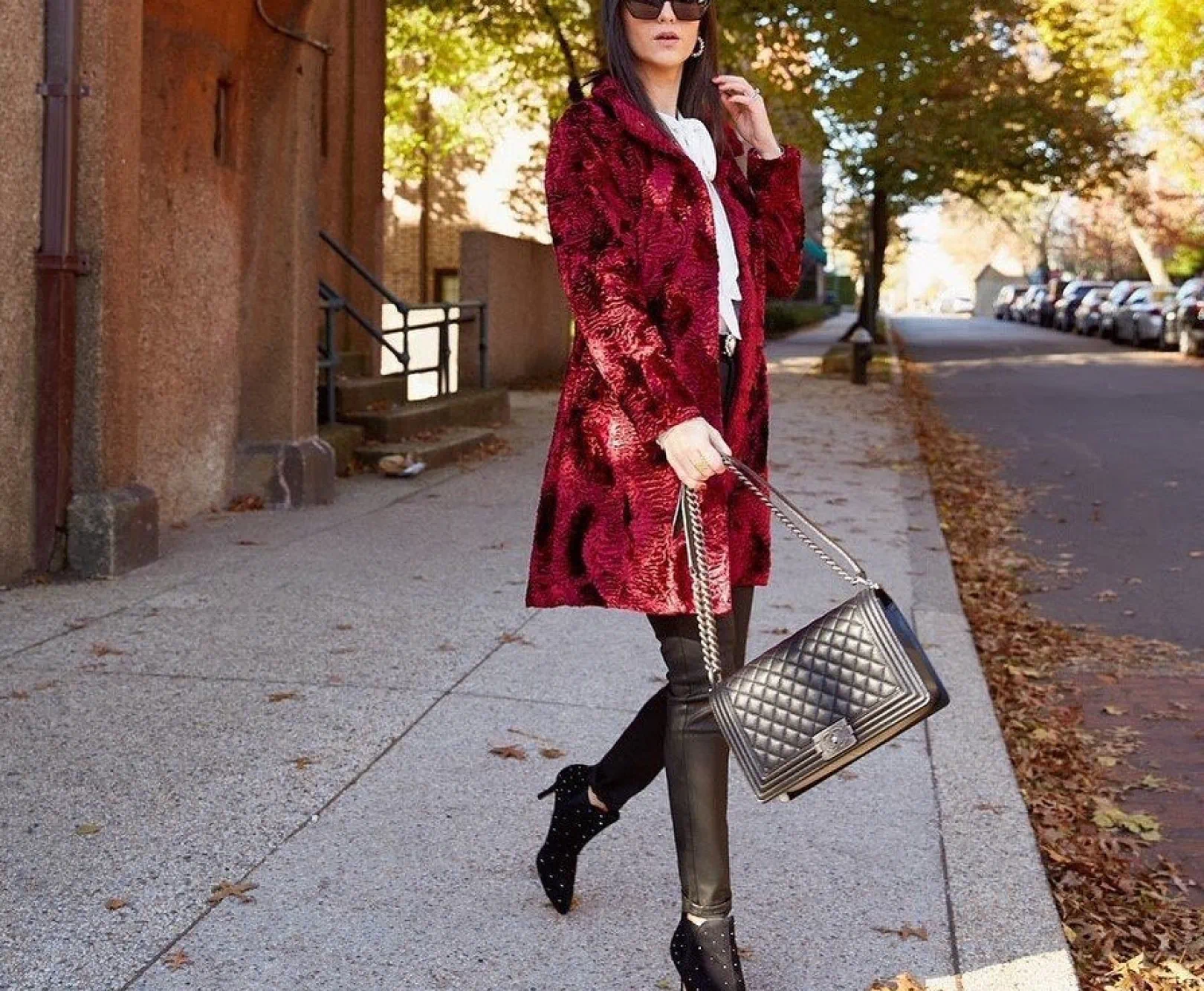 a woman in sunglasses, a red coat, and heels walks along the sidewalk with a luxurious black purse in hand