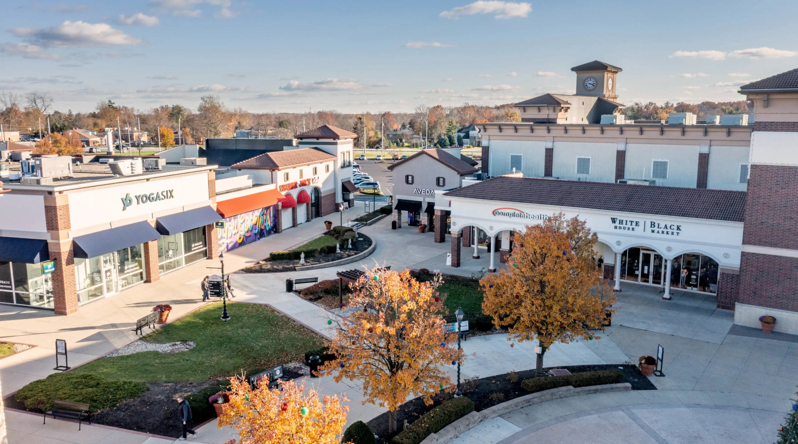 Storefronts in the Jefferson Pointe shopping center, small community area with trees and grass, storefronts including "Trader Joe's", "Aveda", and "Complete Health".