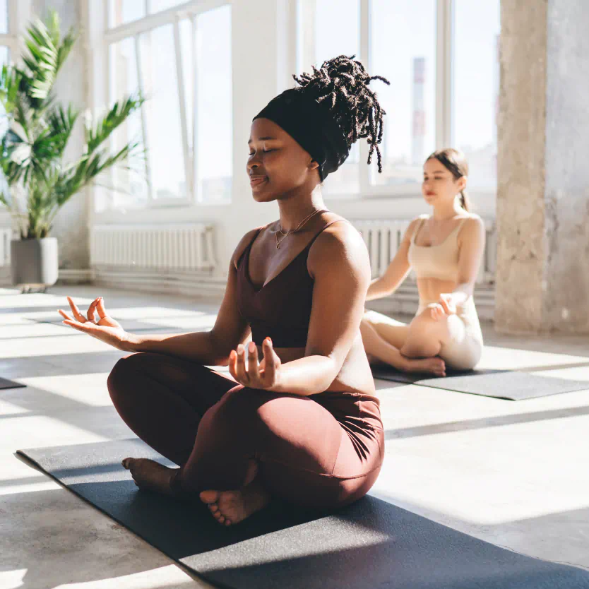 Two people sit cross legged on yoga mat in a well lit yoga studio.