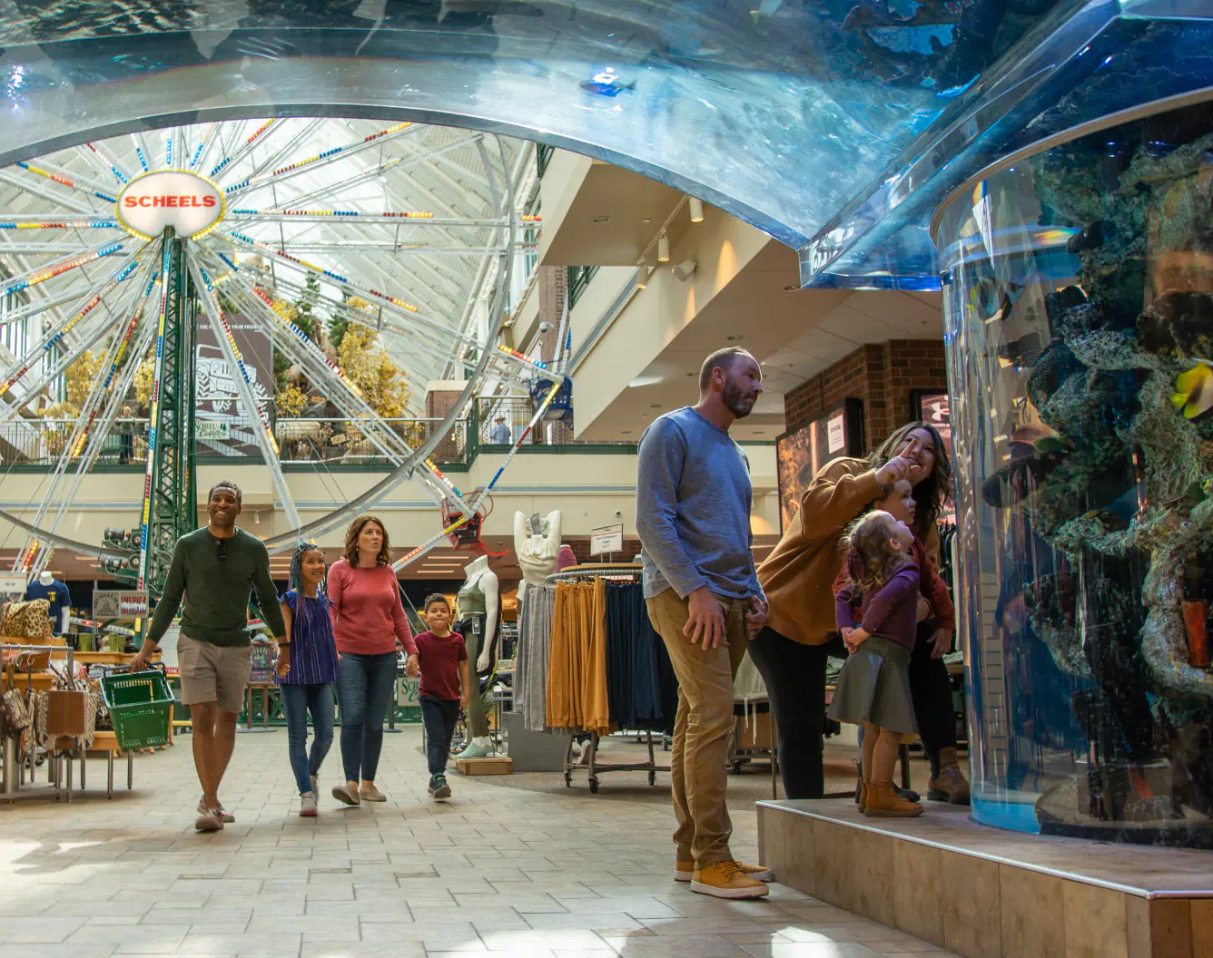Families walk through a great indoor mall space. A ferris wheel fills the space in the background and the families look at a large fish tank in the foreground.