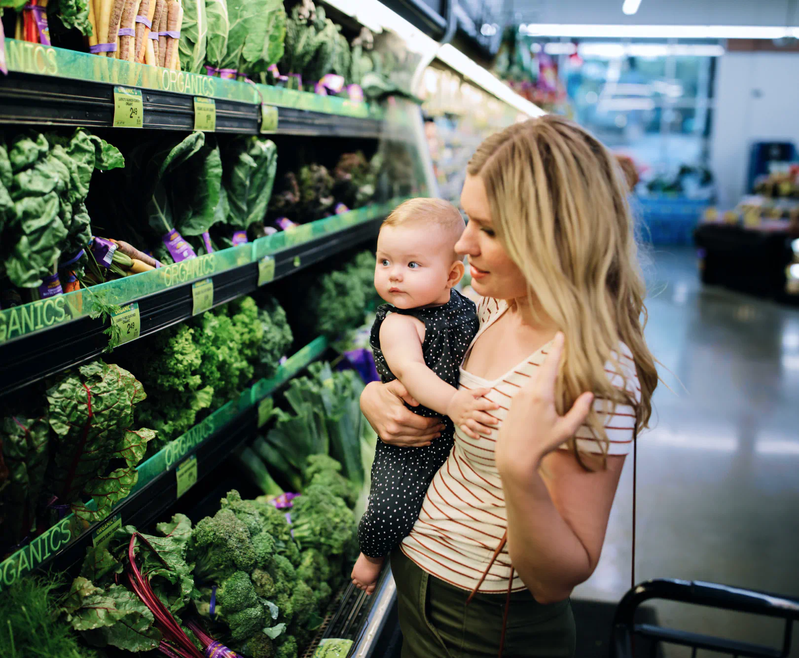 Woman holding her baby while shopping in a grocery store produce aisle