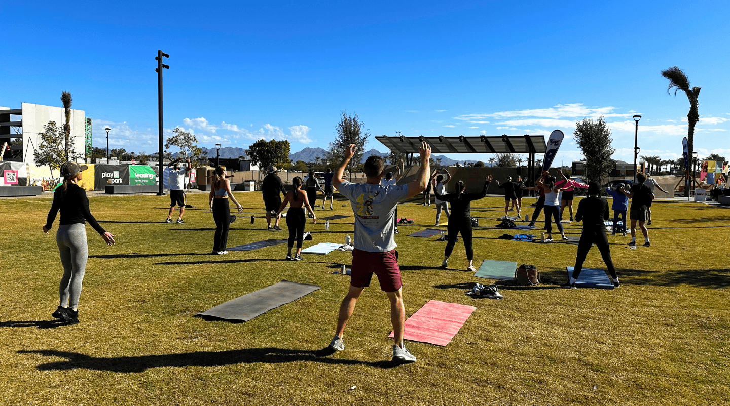 Many people practice yoga on a big lawn. Lots of mats are arranged in rows leading up to an instructor facing the group.
