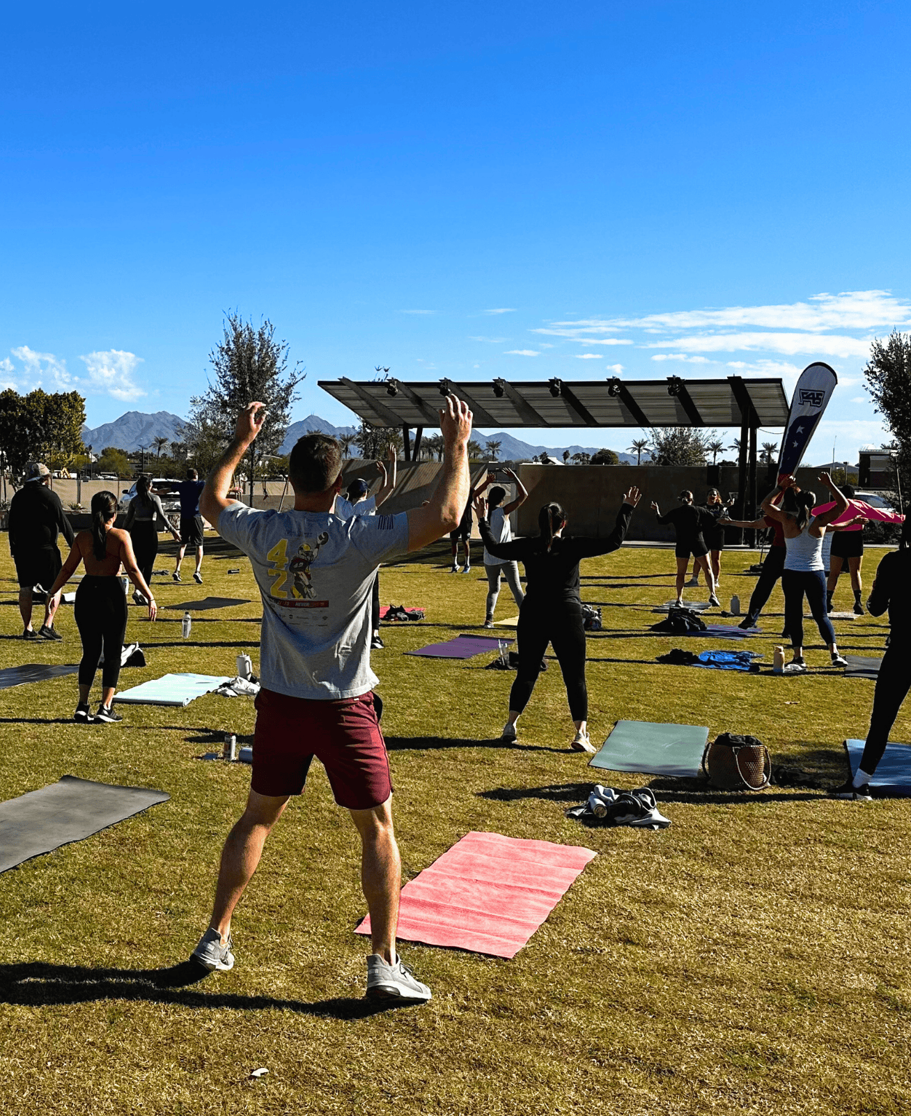 Many people practice yoga on a big lawn. Lots of mats are arranged in rows leading up to an instructor facing the group.