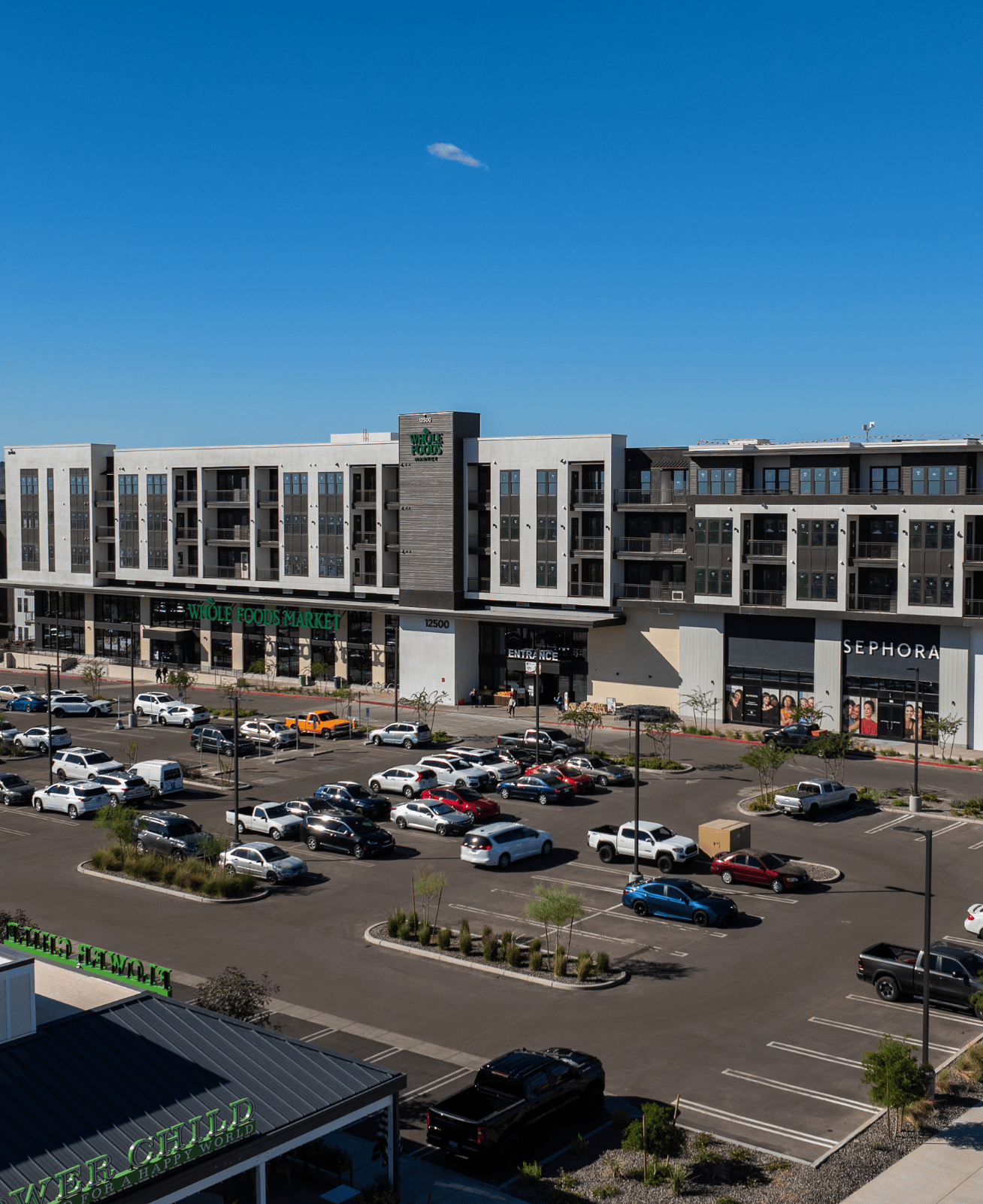 Entrance to shops and the parking lot at Paradise Valley. Many cars parked in front of the storefronts showing "Whole Foods Market", "Sephora", and "Flower Child".
