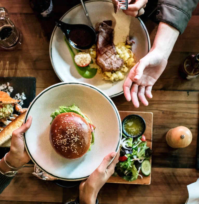 Overhead view of a wooden table filled with plates of food, including a steak and salad. One person hands a hamburger to another person across the table.