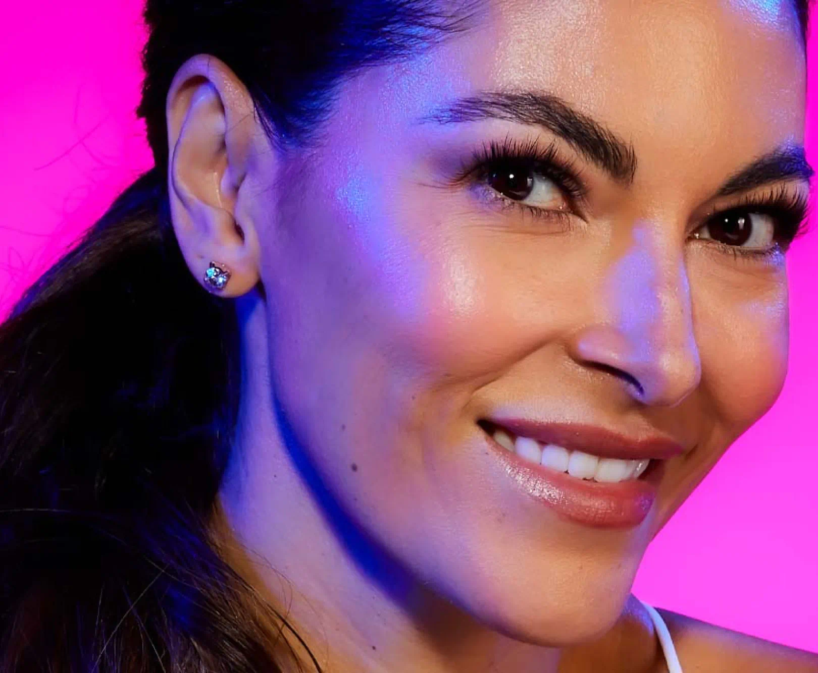 Close-up of a smiling person with dark hair pulled back and wearing a stud earring, set against a pink and purple background.
