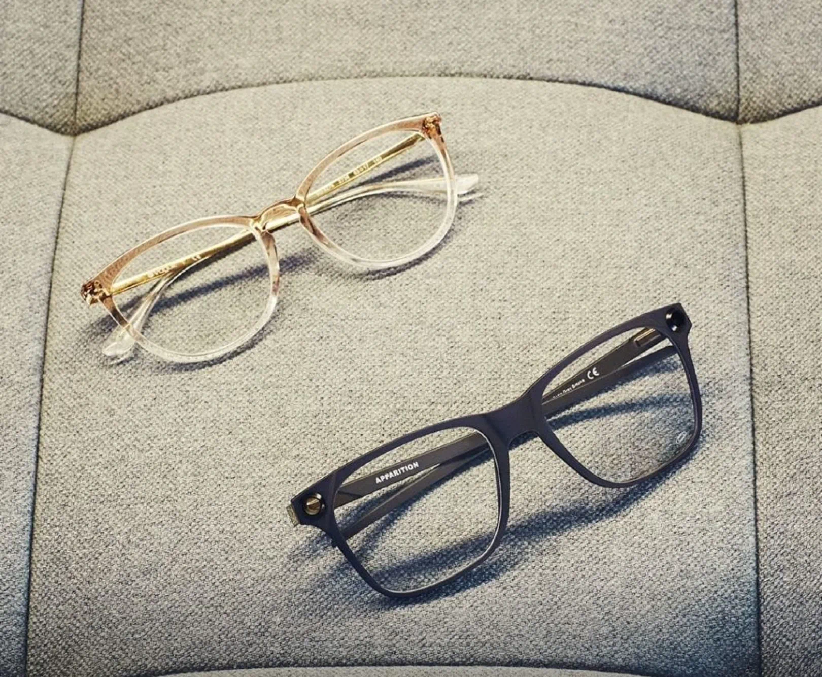 two pairs of glasses, one gold, one black, sit on an upholstered surface