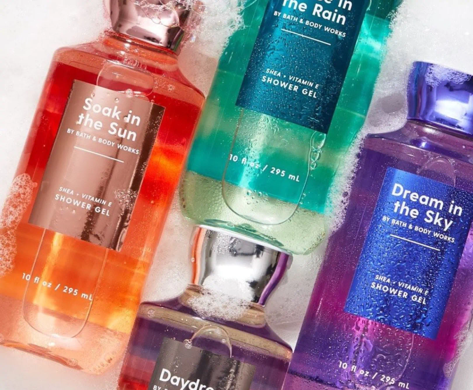 Four colorful Bath & Body Works shower gel bottles on a white frothy surface, each with a metallic cap.