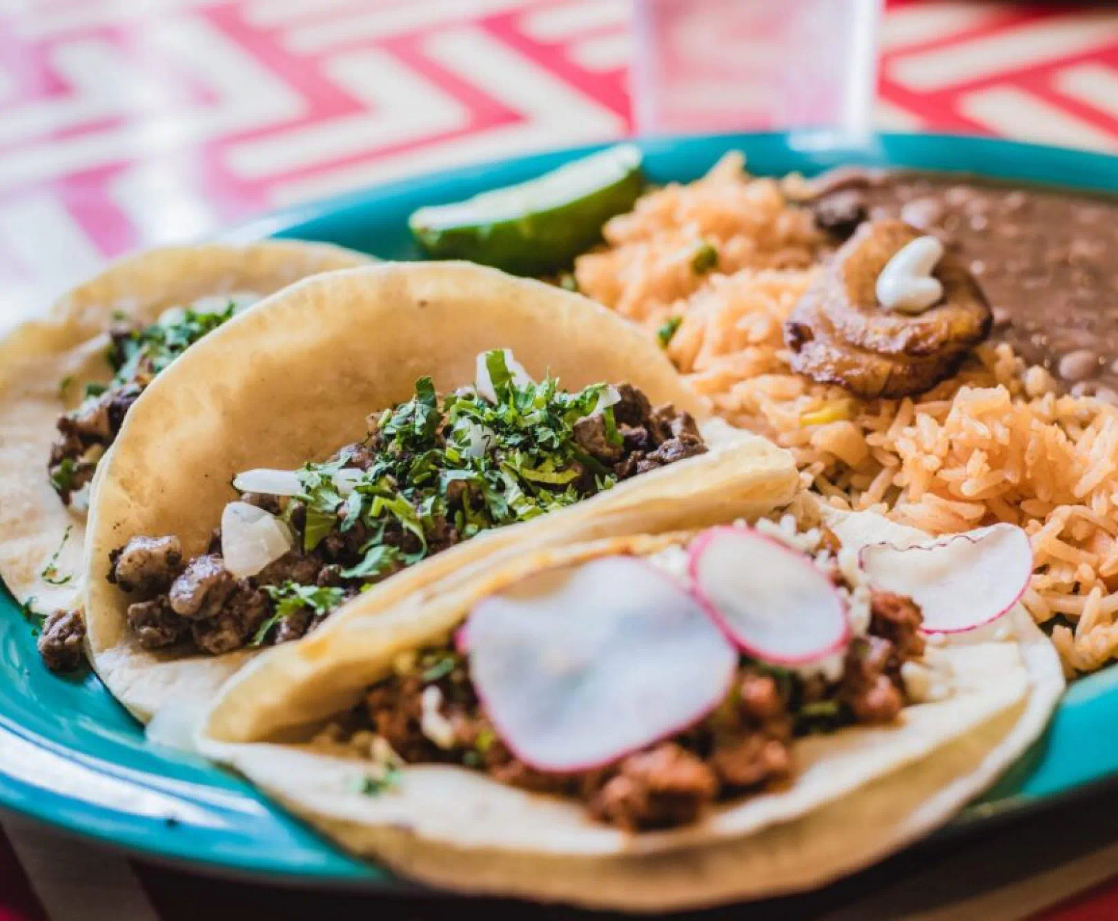 Three tacos with cilantro and onion on a turquoise plate, served with rice, beans, plantains, and a lime wedge.