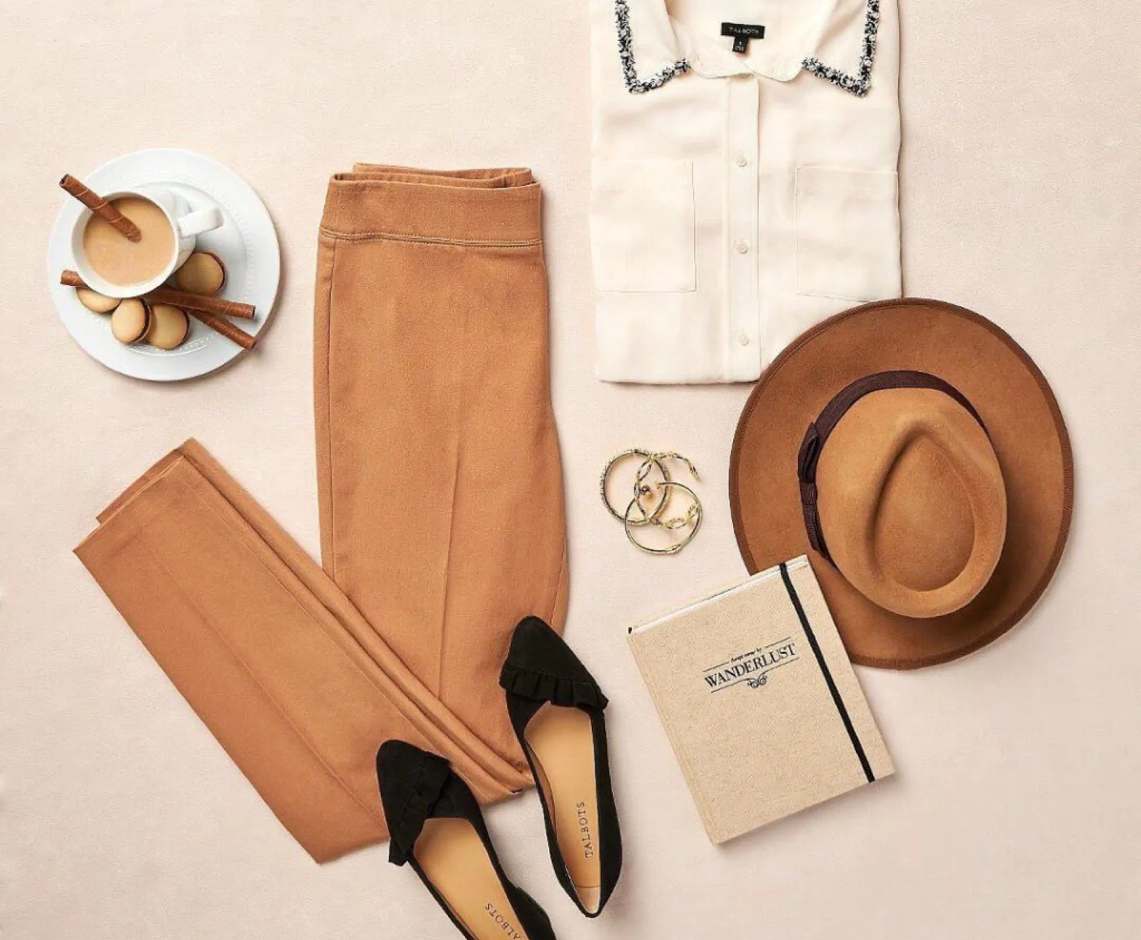 Flat lay of beige trousers, cream blouse, black flats, brown hat, gold bracelets, "Wanderlust" journal, and coffee with cookies.