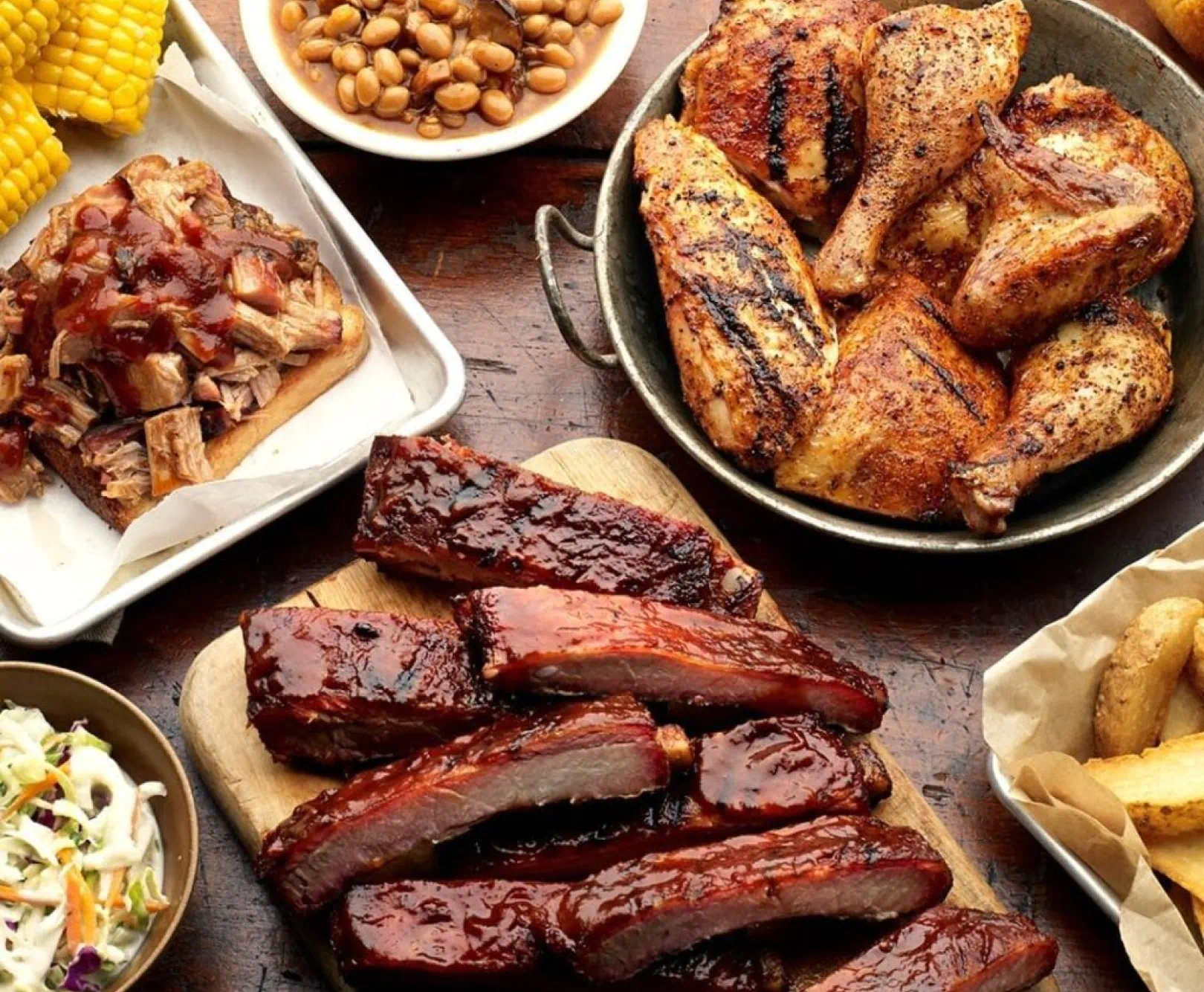 An array of barbecued ribs, grilled chicken, pulled pork, baked beans, corn, coleslaw, and fries on a wooden table.